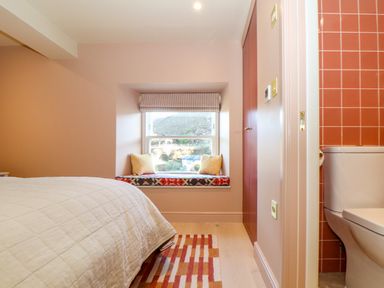 A bedroom with a bed a window seat with cushions a striped rug and part of a bathroom with orange tiles at Mavericks Wave in St Agnes