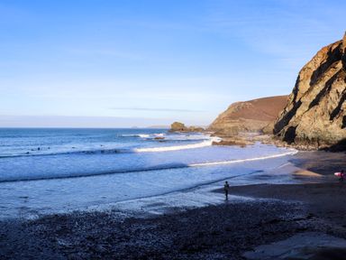 A beach with surfers in the water and rocky cliffs in the background at Mavericks Wave in St Agnes
