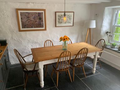 A dining room with a wooden table and six chairs with cushions a vase of sunflowers on the table two framed pictures on the wall a floor lamp and a window with plants on the sill at Kernyk Carn Brea near Pool