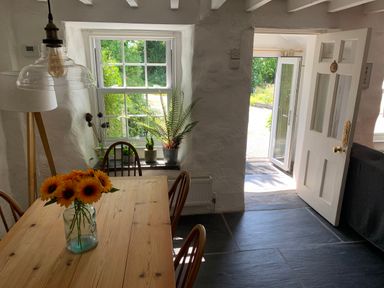 A dining room with a wooden table and sunflowers in a vase near a window and an open door at Kernyk in Carn Brea near Pool