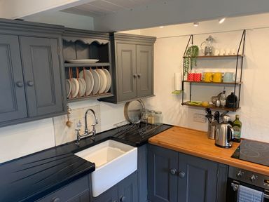 A kitchen corner with grey cabinets a white sink dishes drying rack and shelves with mugs and kitchen items at Kernyk Carn Brea near Pool