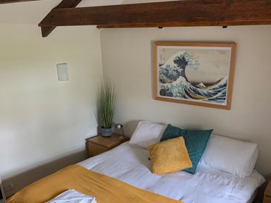 A bedroom with a bed with white sheets and yellow and green pillows and a wooden nightstand with a plant and lamp and a framed wave artwork on the wall at Kernyk Carn Brea near Pool