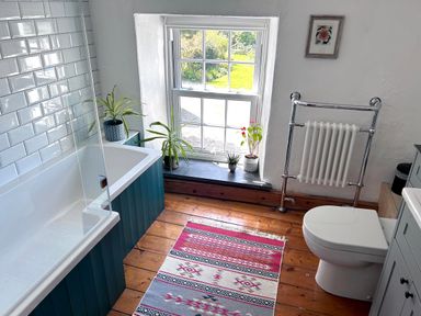 A bathroom with a bathtub, toilet, plants on the windowsill, a radiator, and a patterned rug on a wooden floor at Kernyk in Carn Brea near Pool