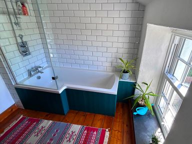 A bathroom with a bathtub and shower next to a window with plants on the windowsill and a colorful rug on the wooden floor at Kernyk in Carn Brea near Pool