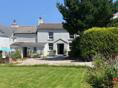 A white two story house with a gravel driveway and a green lawn with shrubs and flowers at Kernyk in Carn Brea near Pool