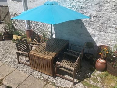 An outdoor patio area with a wooden table and four chairs under a blue umbrella surrounded by plants and pots at Kernyk in Carn Brea near Pool