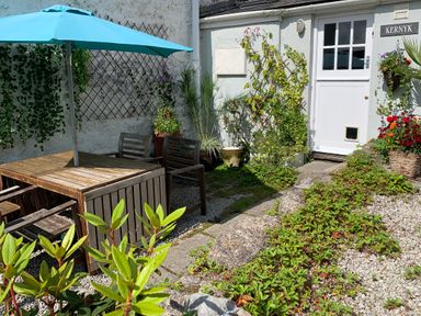 An outdoor patio with wooden table and chairs under a blue umbrella and plants near a white door at Kernyk Carn Brea near Pool