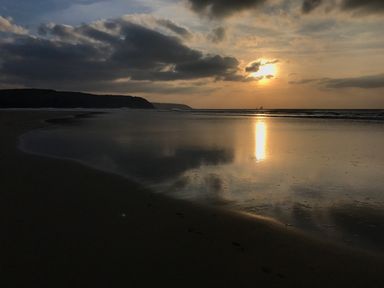 A beach with footprints in the sand and the sun setting over the water at Kernyk in Carn Brea near Pool