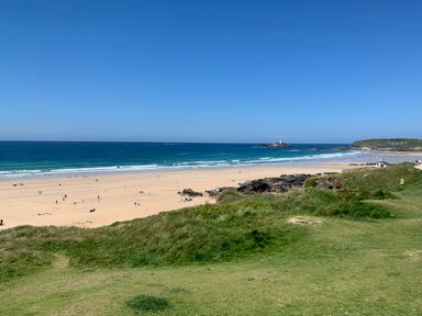 A sandy beach with people scattered around a grassy hill and a lighthouse on a rocky island at Kernyk in Carn Brea near Pool