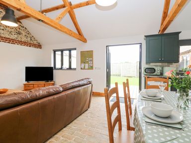 An open living and dining area with wooden beams a leather sofa a TV on a wooden stand a dining table set with dishes and flowers at The Old Dairy in Tittleshall near Brisley