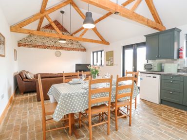 A dining area with a table set for six and wooden chairs next to a kitchen with green cabinets and a living area with a brown sofa at The Old Dairy in Tittleshall near Brisley