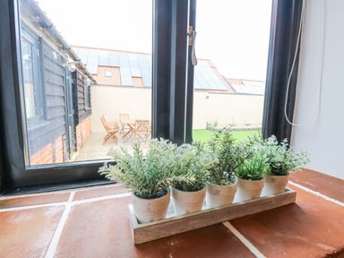 A window with five small potted plants on the windowsill overlooking an outdoor patio with wooden chairs and a table at The Old Dairy in Tittleshall near Brisley
