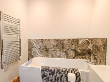 A bathroom with a bathtub featuring brown stone tiles a patterned towel a toilet paper holder and a chrome heated towel rail at The Old Dairy in Tittleshall near Brisley
