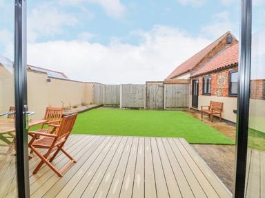 A backyard with a wooden deck table and chairs artificial grass and a brick building at The Old Dairy in Tittleshall near Brisley