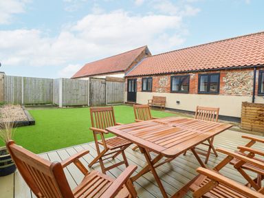 An outdoor patio with wooden table and chairs next to a lawn and a brick building with tiled roof at The Old Dairy in Tittleshall near Brisley