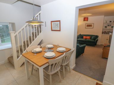 A dining area with a wooden table set for four and beige chairs next to carpeted stairs and an adjoining living room with green sofas at Wood Cottage in Solva