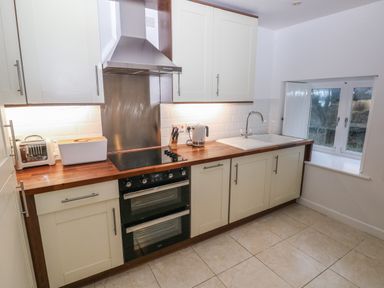 A kitchen with white cabinets a wooden countertop a toaster kettle stove oven and a sink under a window at Wood Cottage in Solva