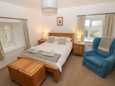 A bedroom with a wooden bed and bedside tables with lamps a blue armchair and windows with curtains at Wood Cottage in Solva