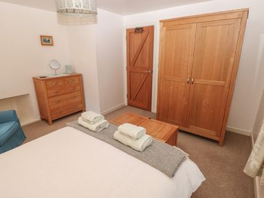 A bedroom with a bed and folded towels a wooden wardrobe a chest of drawers a blue armchair and a wooden door at Wood Cottage in Solva