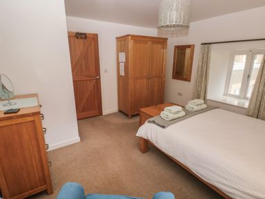 A bedroom with a bed, wooden wardrobe, wooden door, dresser with mirror, and window with curtains at Wood Cottage in Solva