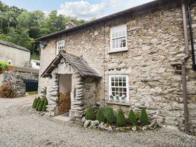 A stone cottage exterior with a wood storage area and small shrubs at Pear Tree Cottage in Grange-Over-Sands