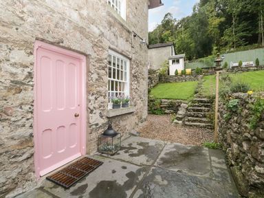 A stone house with a pink door and window with flowers outside a garden with stone steps and green grass at Pear Tree Cottage in Grange-Over-Sands