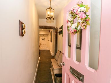 A hallway with a pink front door and floral wreath and a wooden bench with cushions at Pear Tree Cottage in Grange Over Sands