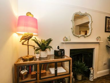 A sideboard with a flamingo lamp and plants next to a fireplace with a mirror above at Pear Tree Cottage in Grange-Over-Sands