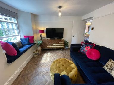 A living room with a blue sofa pink cushions a window seat a yellow ottoman and a TV cabinet at Pear Tree Cottage in Grange-Over-Sands