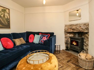 A living room with a blue sectional sofa with colorful cushions a yellow ottoman a fireplace with a stone surround a basket of firewood and a mirror above the fireplace at Pear Tree Cottage in Grange-Over-Sands