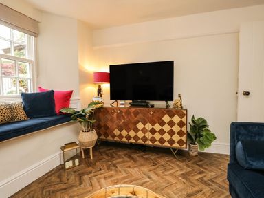A living room with a television on a patterned wooden cabinet and a window seat with colorful cushions at Pear Tree Cottage in Grange-Over-Sands
