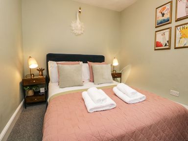 A bedroom with a bed with pink quilt and pillows two bedside tables with lamps and wall art at Pear Tree Cottage in Grange-Over-Sands