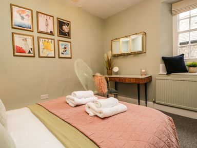 A bedroom with a bed linens, framed pictures on a wall, a vanity table with a mirror and a chair, and a window seat with cushions at Pear Tree Cottage in Grange-Over-Sands