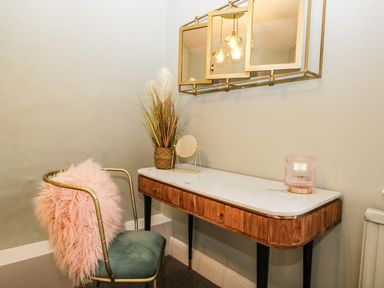 A dressing table with a mirror light and vase next to a chair with a pink cushion at Pear Tree Cottage in Grange-Over-Sands