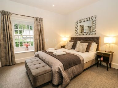 A bedroom with a bed with pillows and folded towels a mirror above the bed bedside tables with lamps a window with curtains and an ottoman at Pear Tree Cottage in Grange-Over-Sands