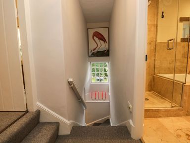 A staircase with gray carpet leading down past a window with pink candles and a flamingo picture above with a bathroom featuring a glass shower door on the right at Pear Tree Cottage in Grange-Over-Sands