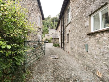 A gravel pathway between stone buildings with a wooden gate and greenery at Pear Tree Cottage in Grange-Over-Sands