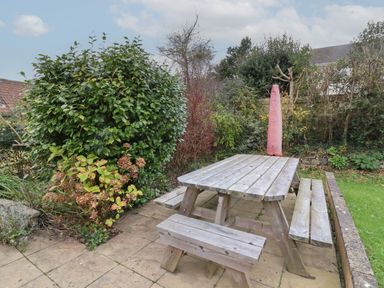 A wooden picnic table with benches on a paved patio surrounded by bushes and greenery at Sunnymead in Bickington near Barnstaple