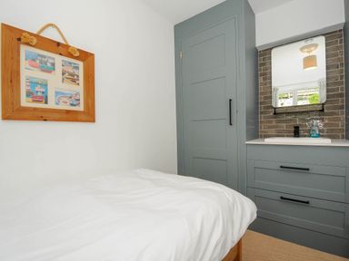 A bedroom with a bed covered in white bedding a framed picture on the wall and a grey wardrobe and sink unit with a mirror at Slipway 14 in Rock