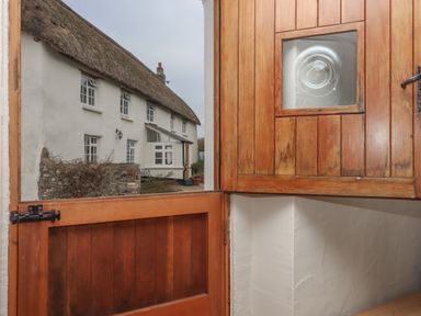 A half-open wooden door with a small glass window showing a thatched roof cottage outside at Cherryblossom Cottage in Winkleigh
