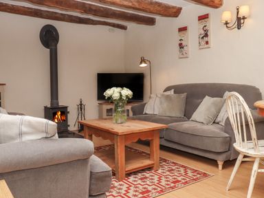 A living room with a wood stove a wooden coffee table with flowers a gray sofa and armchair a television and wooden ceiling beams at Cherryblossom Cottage in Winkleigh