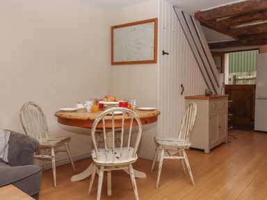 A dining area with a round wooden table set for breakfast with four white distressed wooden chairs near a gray sofa and wooden cabinet with beams on ceiling at Cherryblossom Cottage in Winkleigh