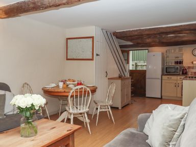 A living area with a wooden dining table and chairs next to a kitchen with a refrigerator and wooden beams on the ceiling at Cherryblossom Cottage in Winkleigh