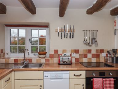 A kitchen with a window above a sink wooden countertop tiled backsplash kitchen knives and utensils hanging on the wall toaster and oven with red checkered towels at Cherryblossom Cottage in Winkleigh