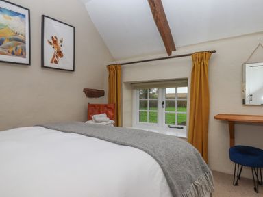 A bedroom with a bed covered in white and grey bedding a window with yellow curtains a chair with towels a wooden desk and two framed pictures on the wall at Cherryblossom Cottage in Winkleigh