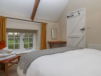 A bedroom with a bed a wooden chair with towels a window with yellow curtains a wooden desk a mirror and a white door at Cherryblossom Cottage in Winkleigh