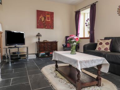A living room with a coffee table with flowers a sofa a TV on a stand and a window with purple curtains at Ty Llugwy in Capel Curig near Betws-Y-Coed