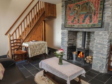 A living room with a stone fireplace and wood burning stove a wooden staircase a table with a floral tablecloth and a chair at Ty Llugwy in Capel Curig near Betws-Y-Coed