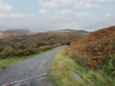 A gravel path curving through grassy hills with scattered bushes and trees under a partly cloudy sky at Ty Siabod in Capel Curig near Betws-Y-Coed