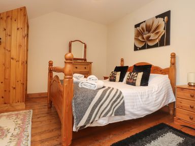A bedroom with a wooden bed with white linens and folded towels a wooden wardrobe dresser and nightstand with a lamp a floral painting on the wall at Ty Siabod in Capel Curig near Betws-Y-Coed